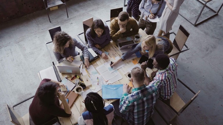 Aerial view of team of employees collaborating on resource plans at large wooden table | SMB resource scheduling