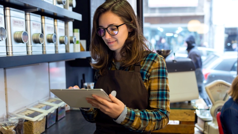 Engaged employee behind retail counter looks at scheduling software using tablet | Reduce Labor Costs