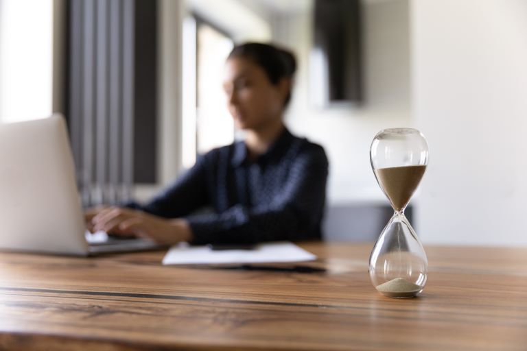 Blurred woman works on laptop computer while hourglass sits on desk in foreground | Improve employee retention